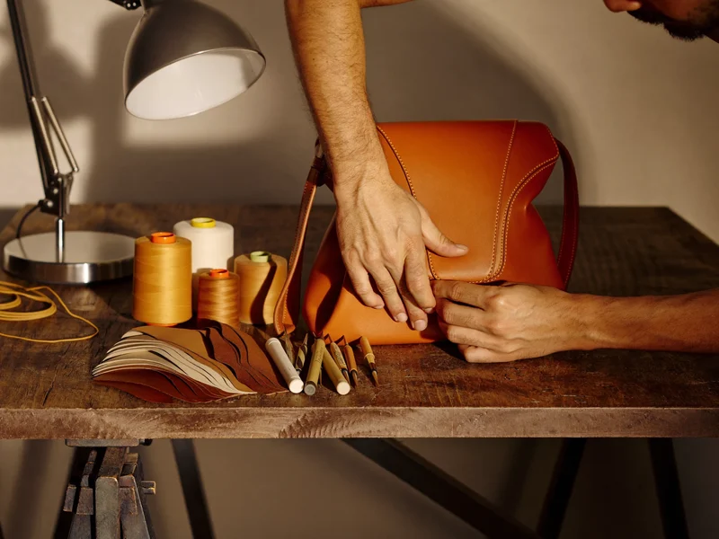 Hände prüfen eine Taschennaht unter einer Lampe auf einer Werkbank