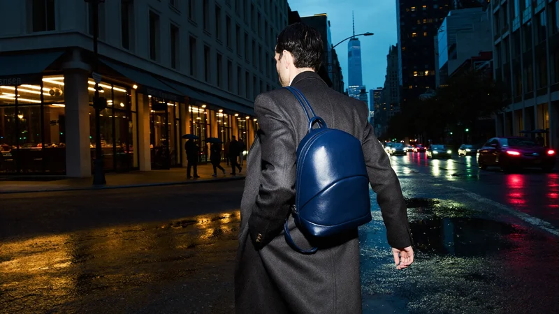 Person geht abends mit Rucksack über der Schulter durch eine regennasse Stadtstraße
